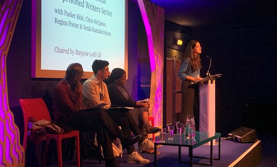 Image shows a woman reading at a lectern whilst four people sit on a stage 