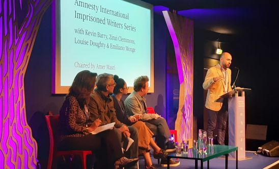 Image shows a man reading at a lectern whilst four people sit on a stage 