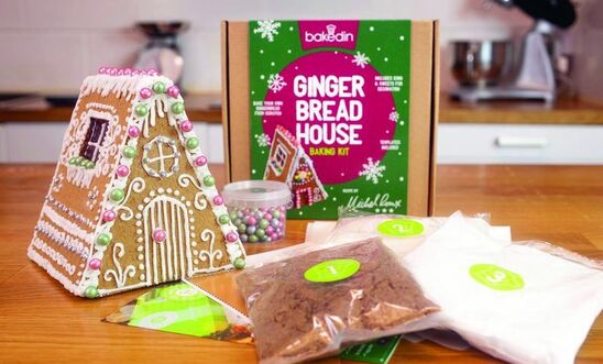 On a kitchen counter is the contents of a gingerbread house kit, including a completed gingerbread house, ingredients and colourful box