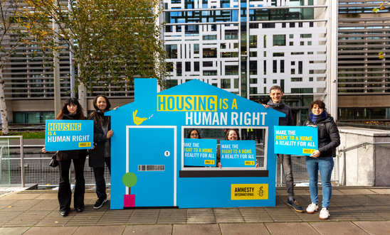 Number of people standing next to a cardboard cutout of a house saying Housing Is a Human Right