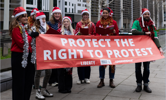 Carol singers wearing festive clothing hold a large red banner saying 'protect the right to protest' outside the Home Office