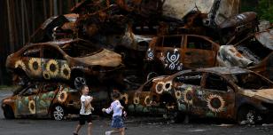 Image shows 2 children playing in front of a pile of burnt cars painted with the Ukrainian national flower, the sunflower 